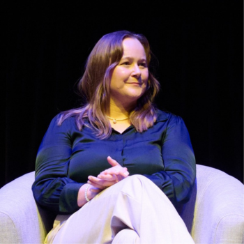A woman seated on stage, smiling with hands clasped, wearing a dark blouse and light trousers.